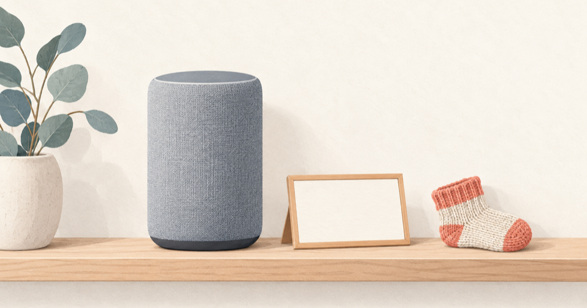 A plain smart speaker on a wooden shelf next to a blank name card and a knitted baby sock